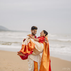 Happy South Indian bride and groom posing in traditional wedding attire on a beach, with the groom lovingly lifting the bride.